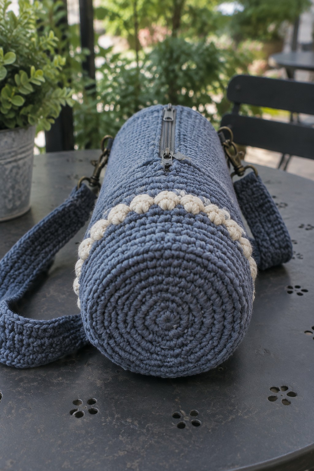 Blue crocheted cylindrical bag with cream bobble trim on dark metal table