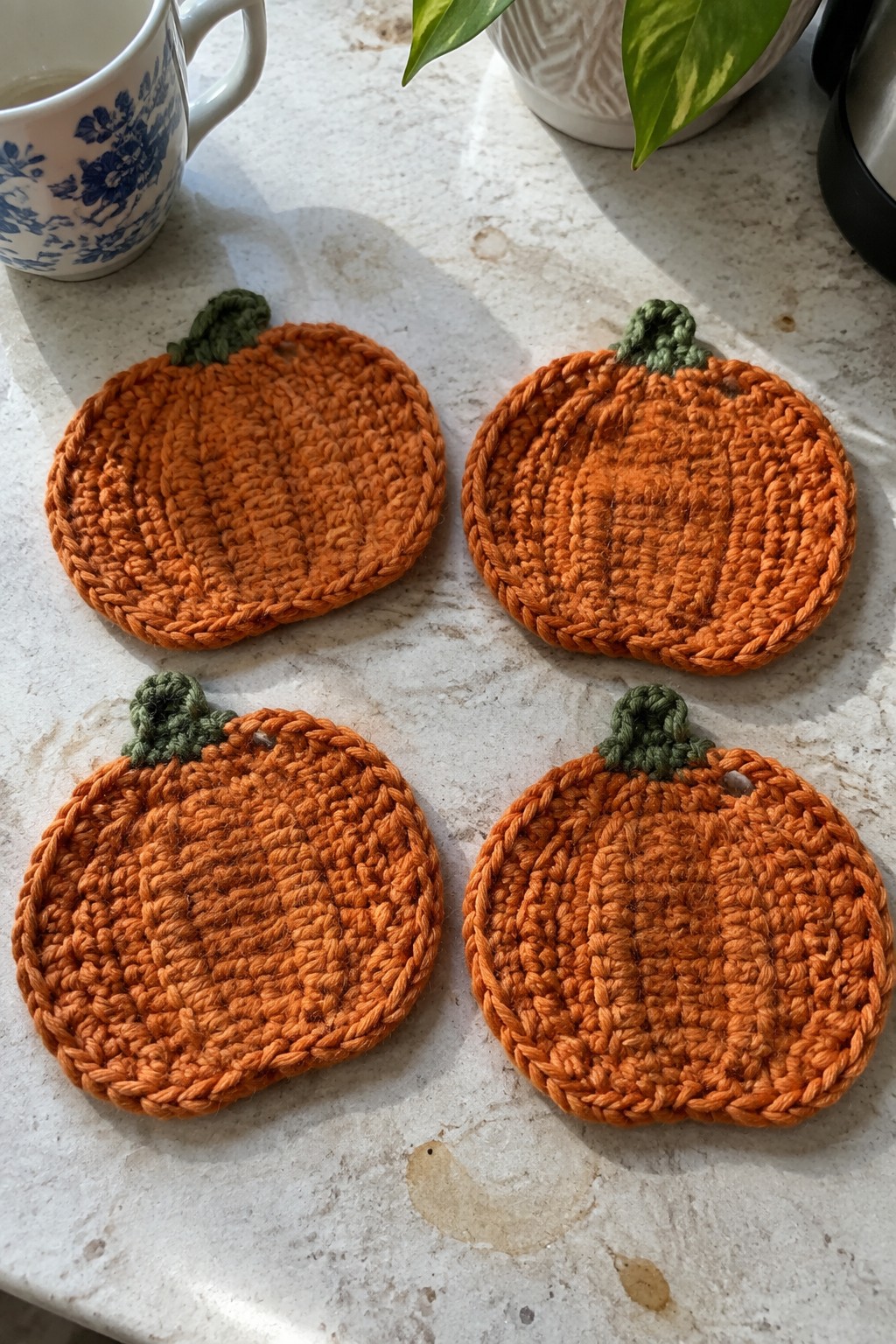 Four orange crocheted pumpkin coasters with green stems on marble countertop