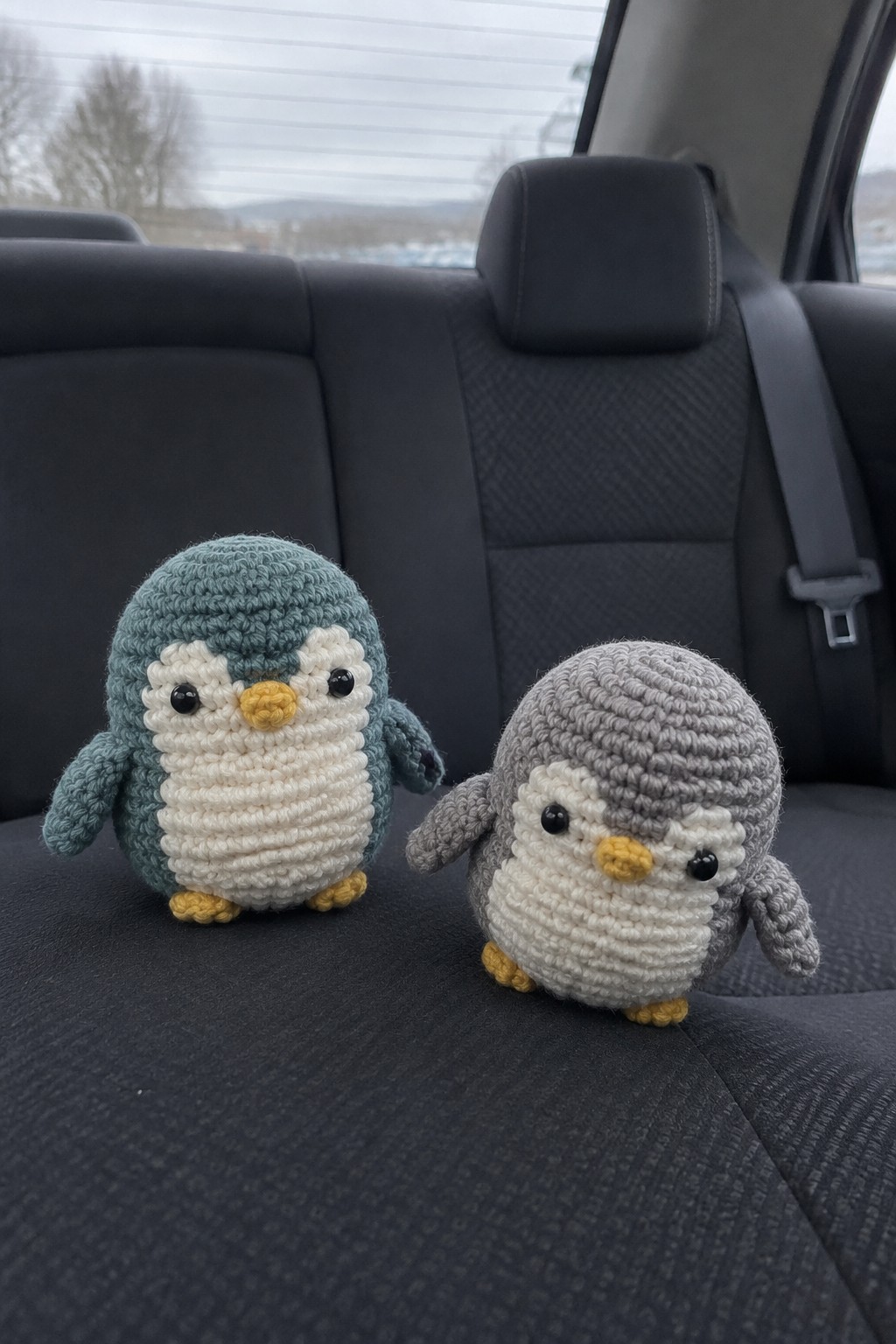 Two crocheted penguins, teal and gray, on a dark car seat.