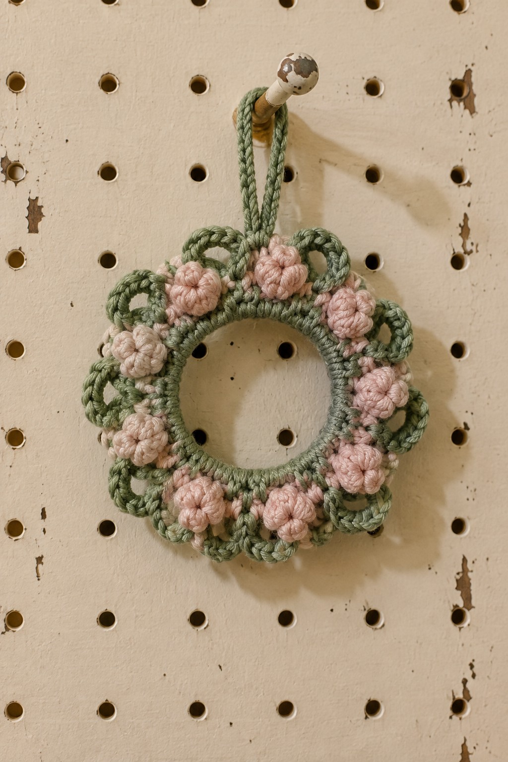 Crocheted green wreath with pink flowers hanging from knob on pegboard.