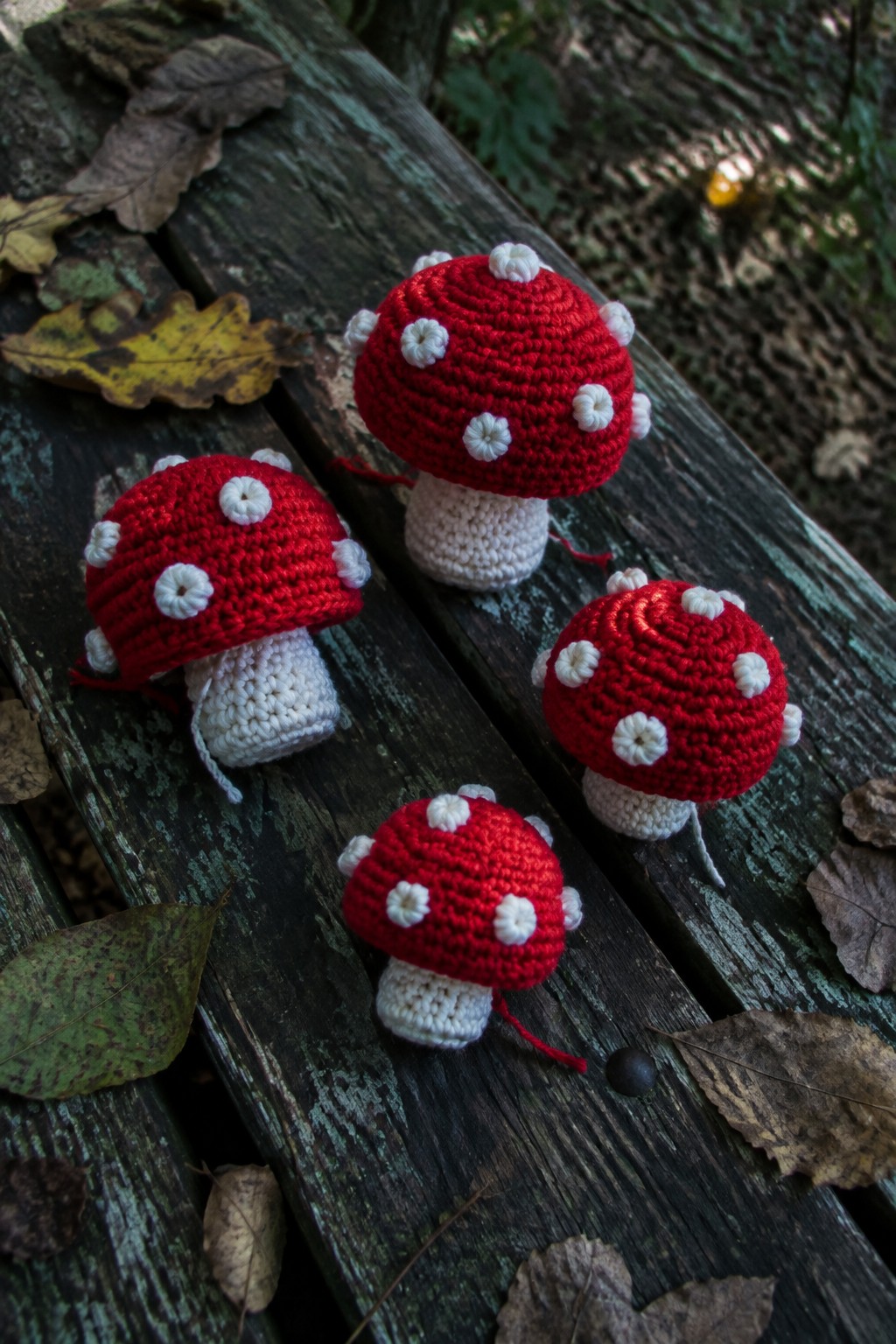 Four crocheted red mushrooms with white spots on a wooden bench amid leaves