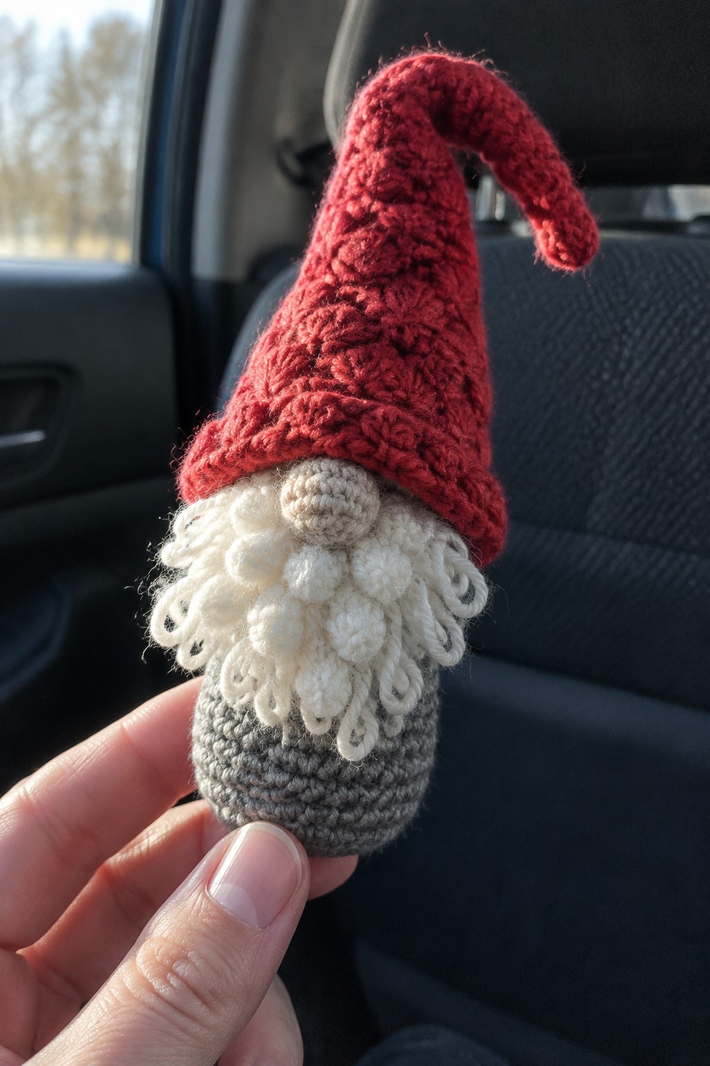 Hand holding crocheted gnome with red hat, white beard, and gray body in car