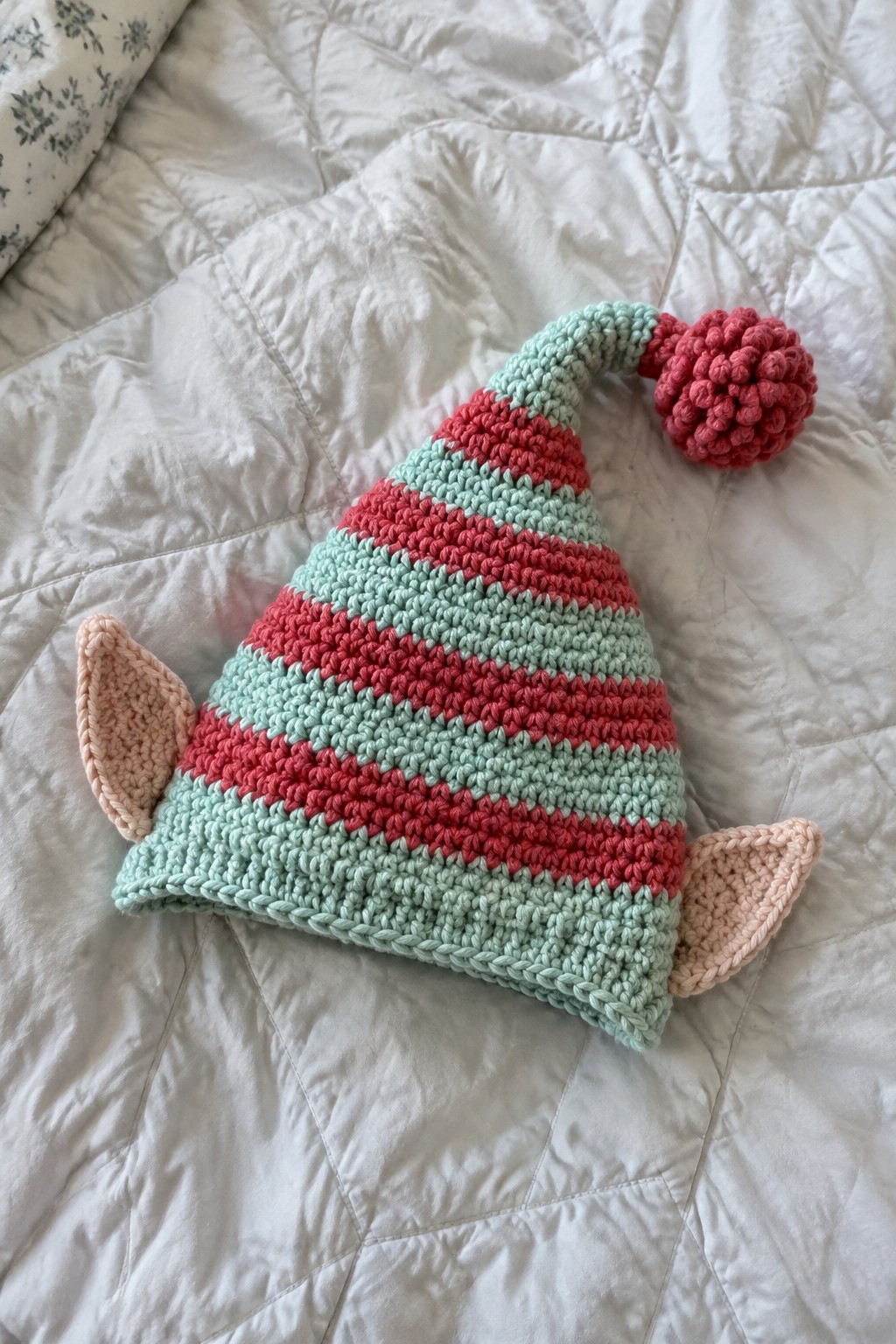 Striped mint and coral crochet hat with ears and pom-pom on white quilt