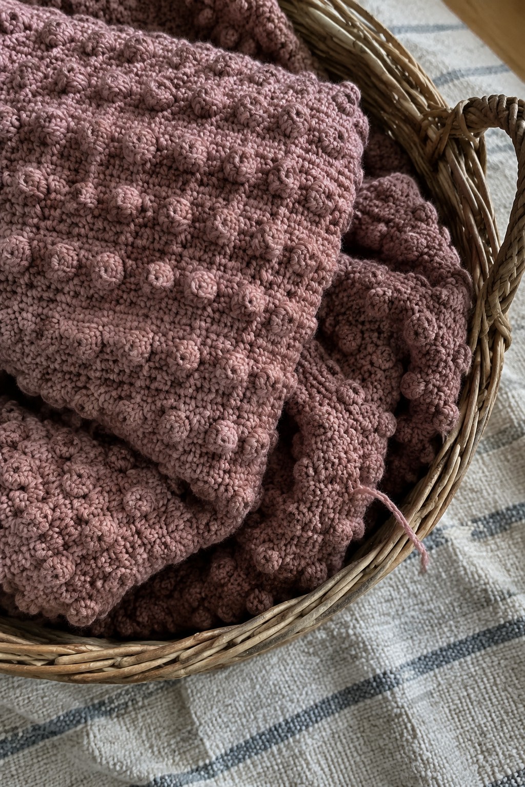 Rose-colored bobble crochet blanket in a wicker basket on striped fabric