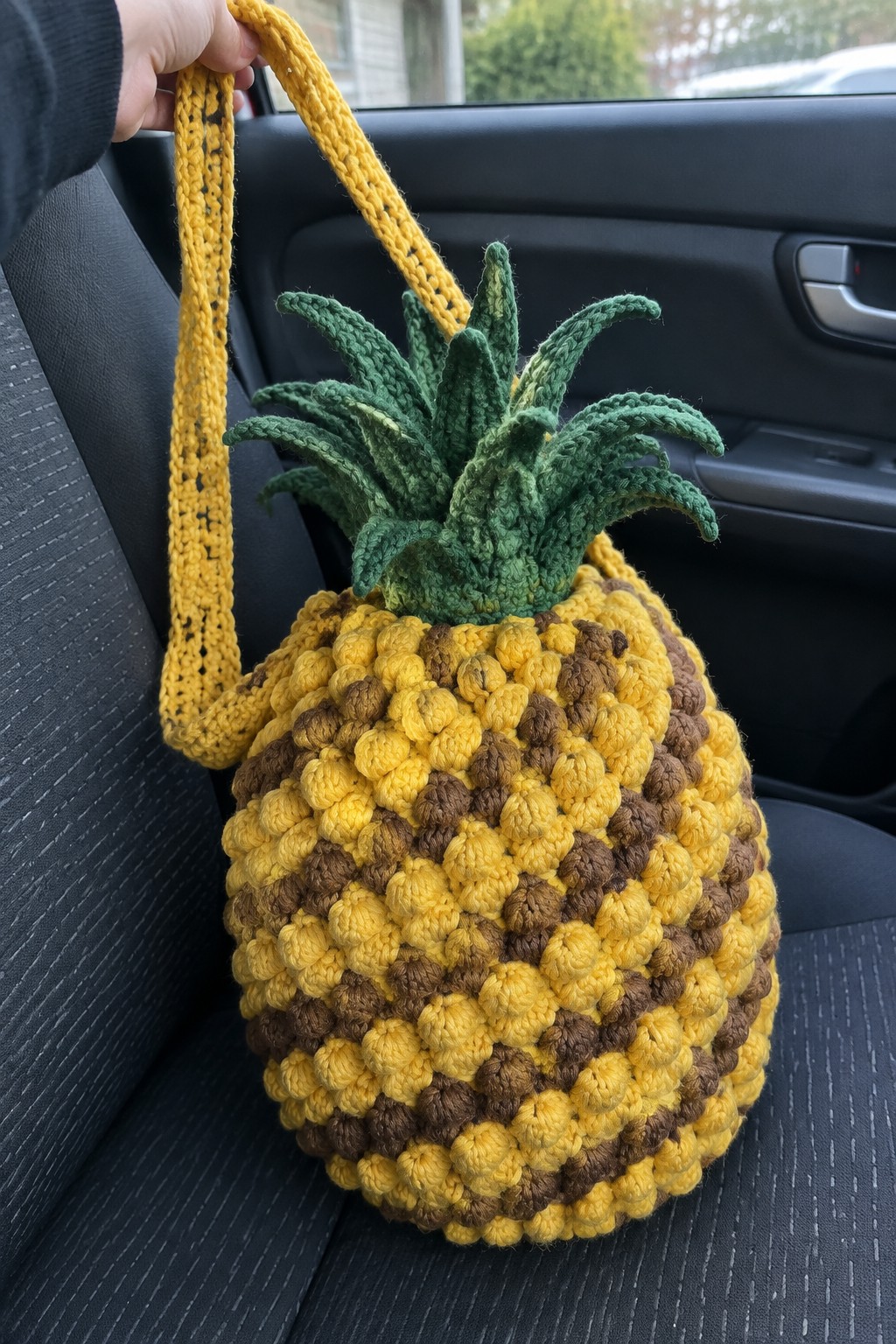 Hand holding crocheted yellow-brown pineapple bag with green top in car