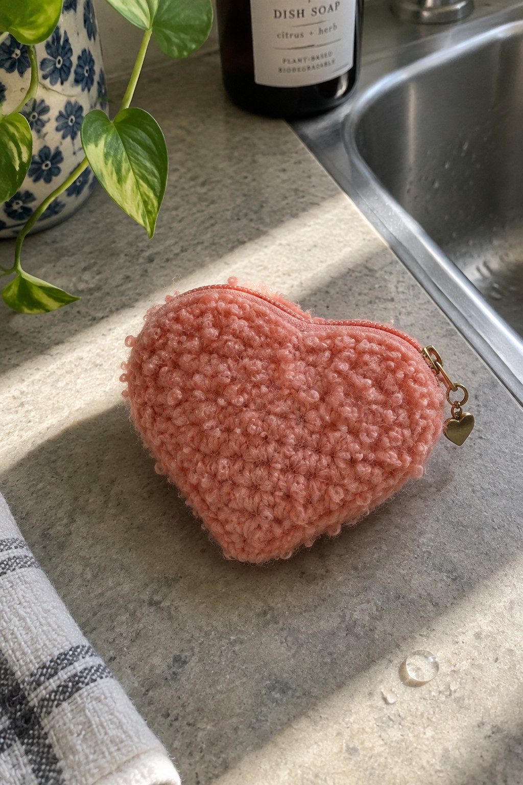 Pink fuzzy heart-shaped zippered pouch with gold charm on kitchen countertop by sink