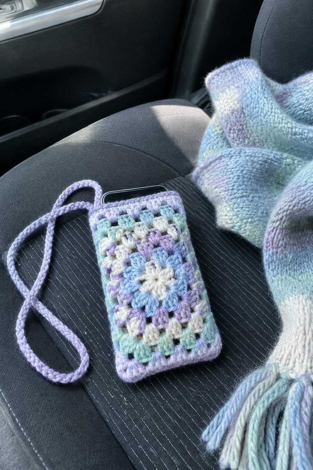 Pastel crocheted phone case with strap on car seat beside knitted scarf