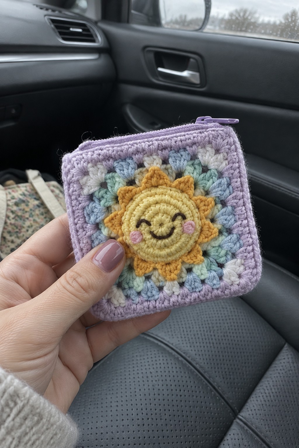 Hand holding pastel crocheted pouch with smiling sun face inside a car