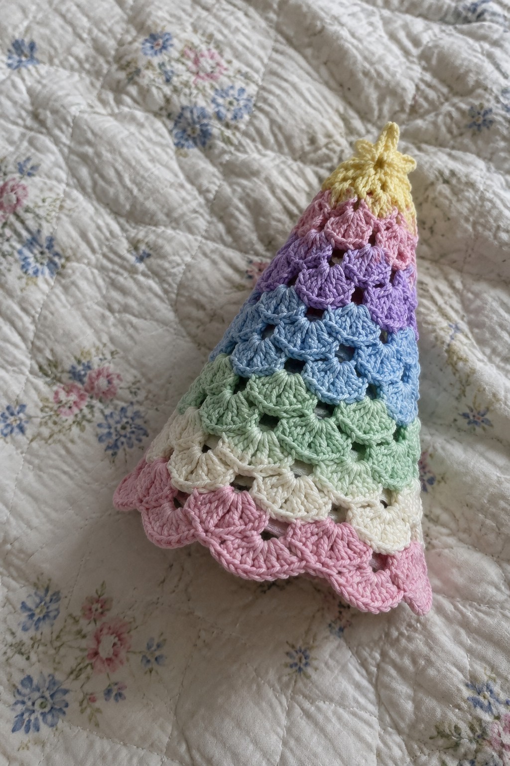 Pastel rainbow crocheted Christmas tree with star topper on floral quilt.