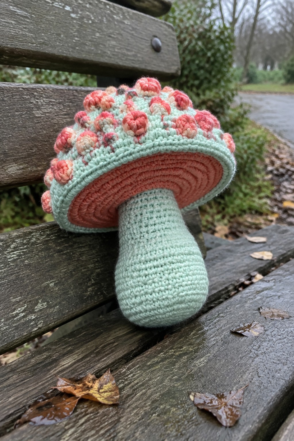 Crocheted mint-green mushroom with pink floral cap on wooden bench with leaves