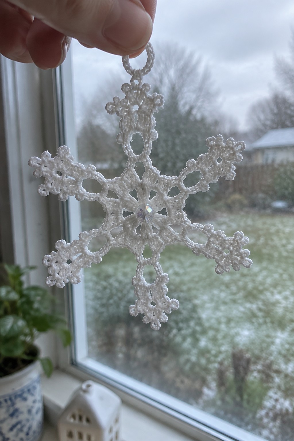 Hand holding white crocheted snowflake ornament with central bead by snowy window