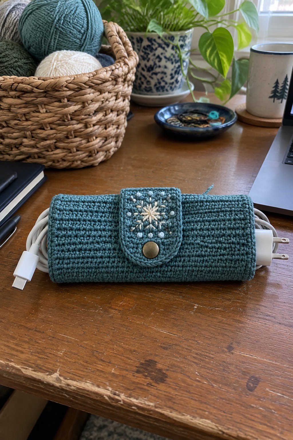 Teal crocheted pouch with snowflake embroidery holding white charging cables on wooden table