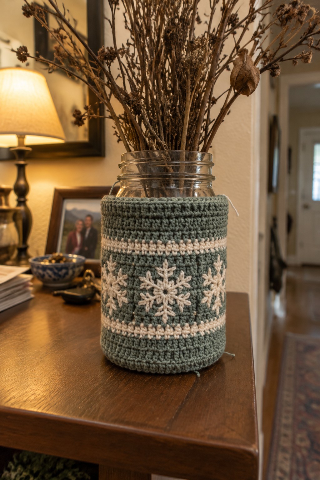 Crocheted green jar cozy with white snowflakes holding dried branches on wooden table.
