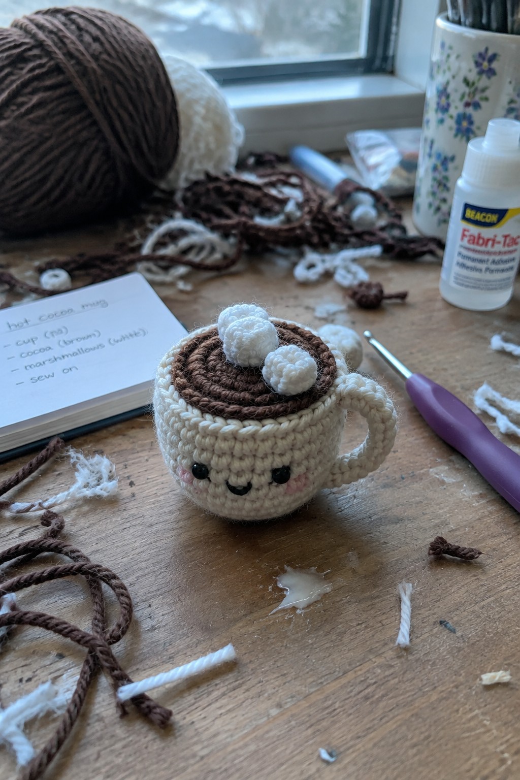 Crocheted hot cocoa mug amigurumi with smiling face and marshmallows on wooden craft table