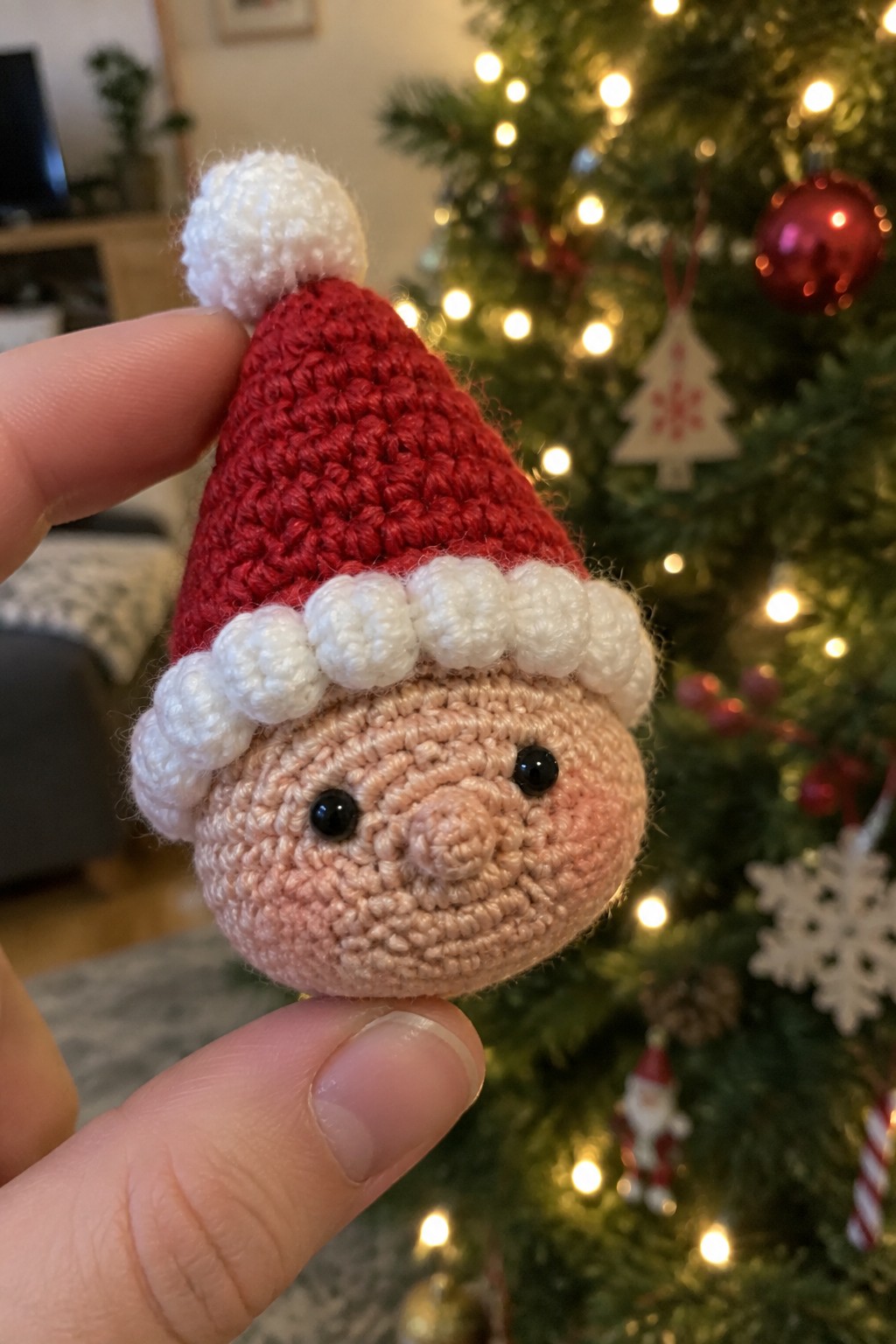 Hand holding crocheted Santa head with red hat and white trim near Christmas tree.