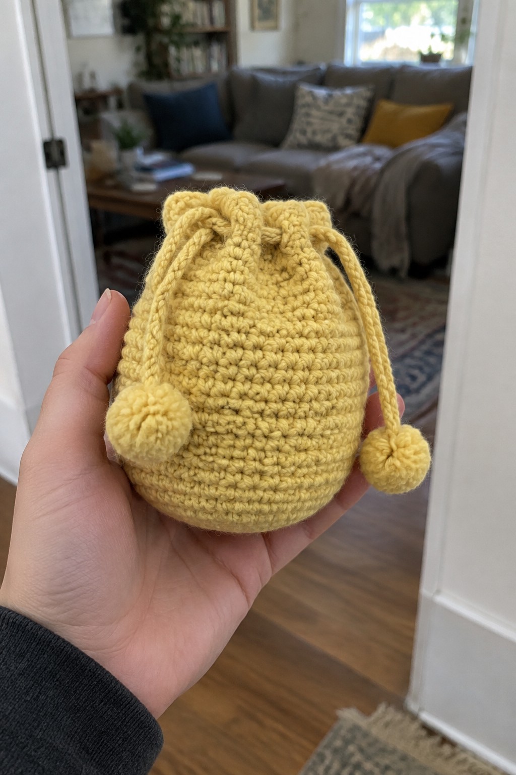 A hand holds a small yellow crocheted drawstring pouch with two pom-poms on the drawstring ends.