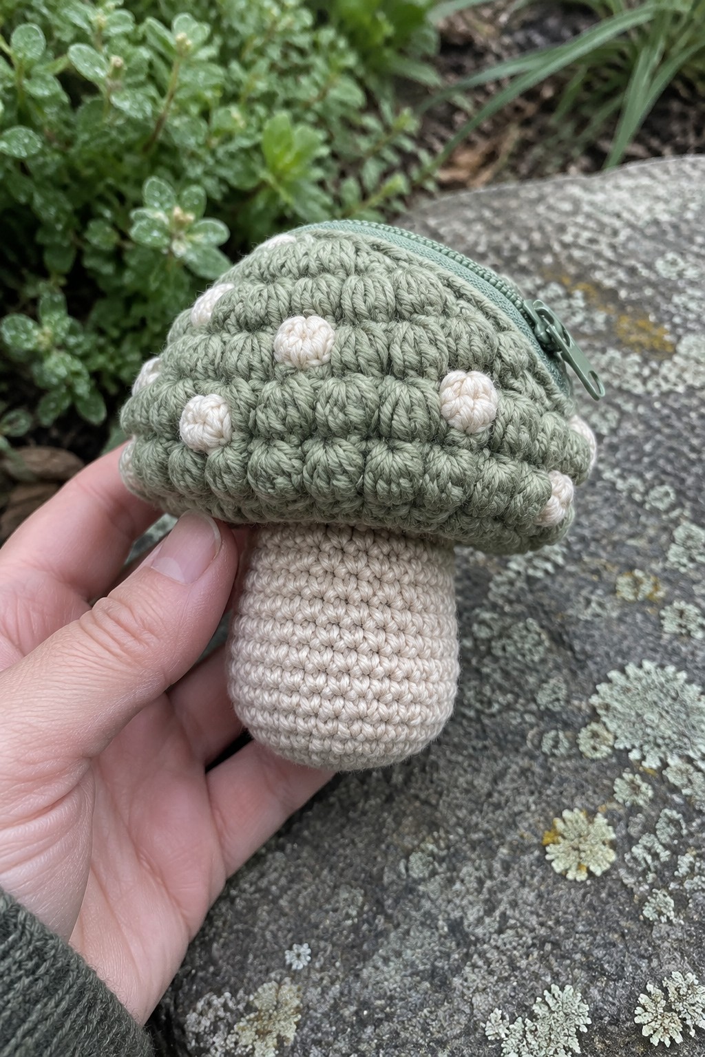 Hand holding sage green crocheted mushroom pouch with white spots and beige stem.