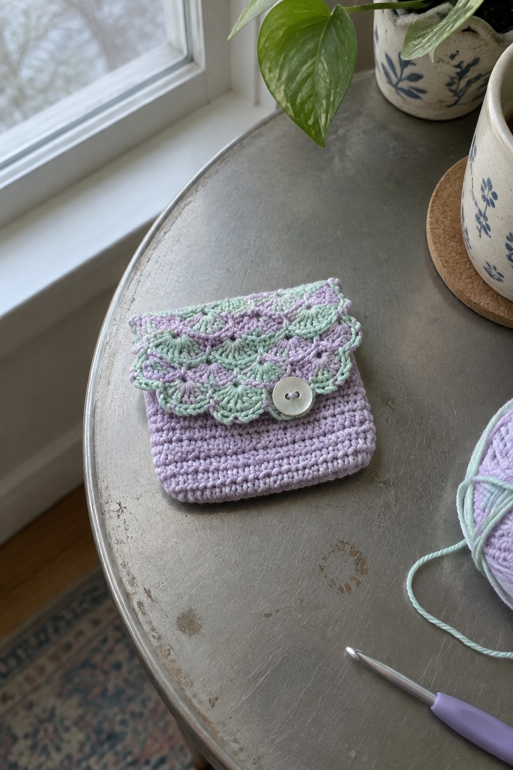 Pastel crocheted pouch with scalloped flap and button on metal table beside hook and yarn