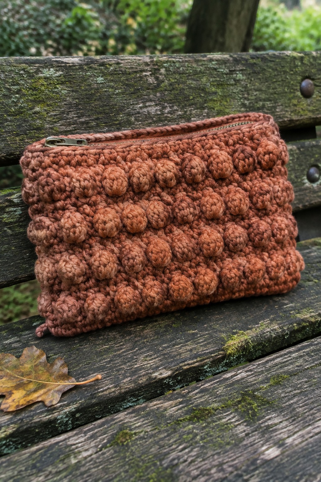 Rust crocheted bobble-stitch pouch with zipper on weathered wooden bench beside dry leaf