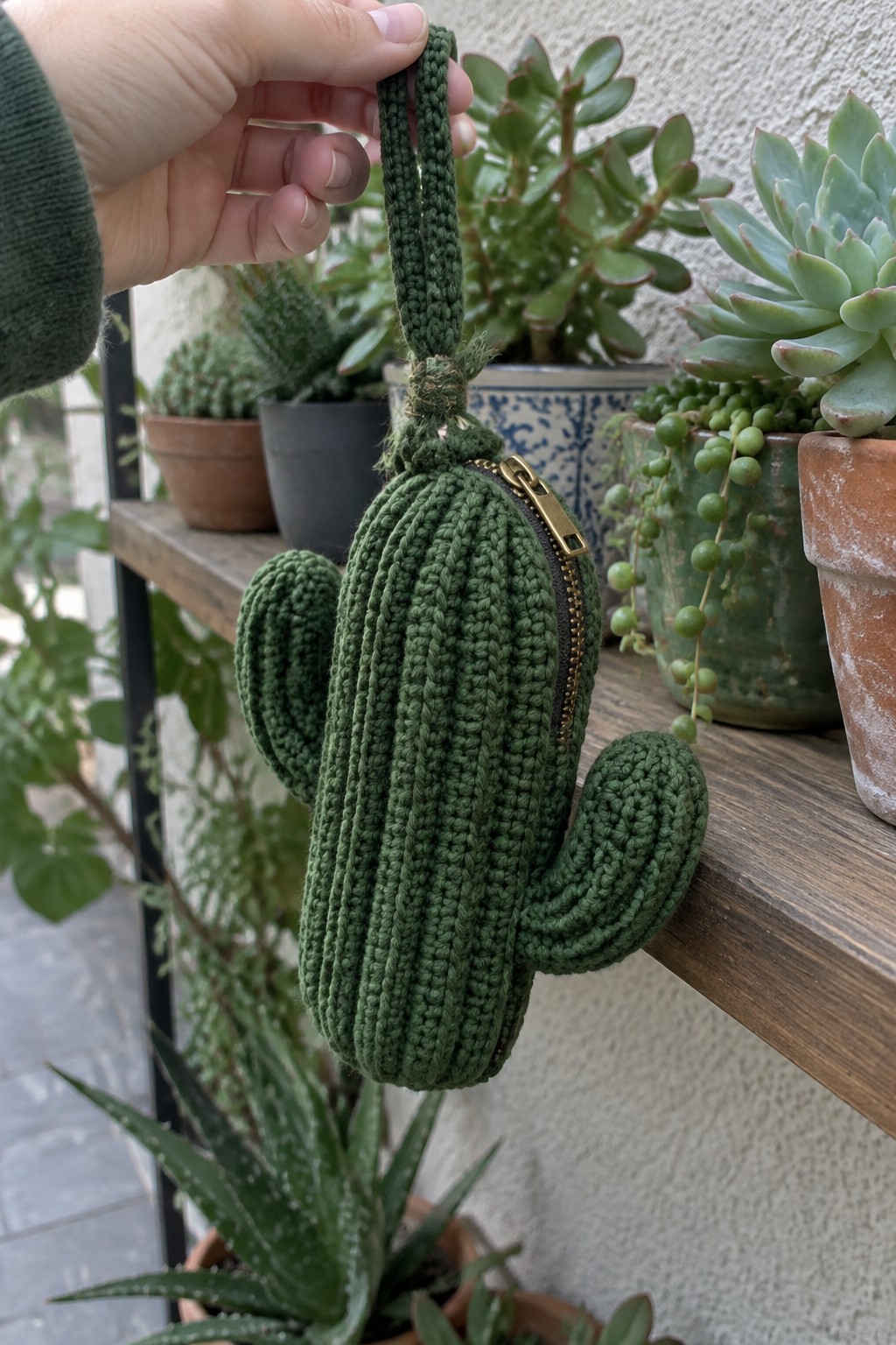 Hand holds green crocheted cactus pouch with zipper amid potted succulents on shelf