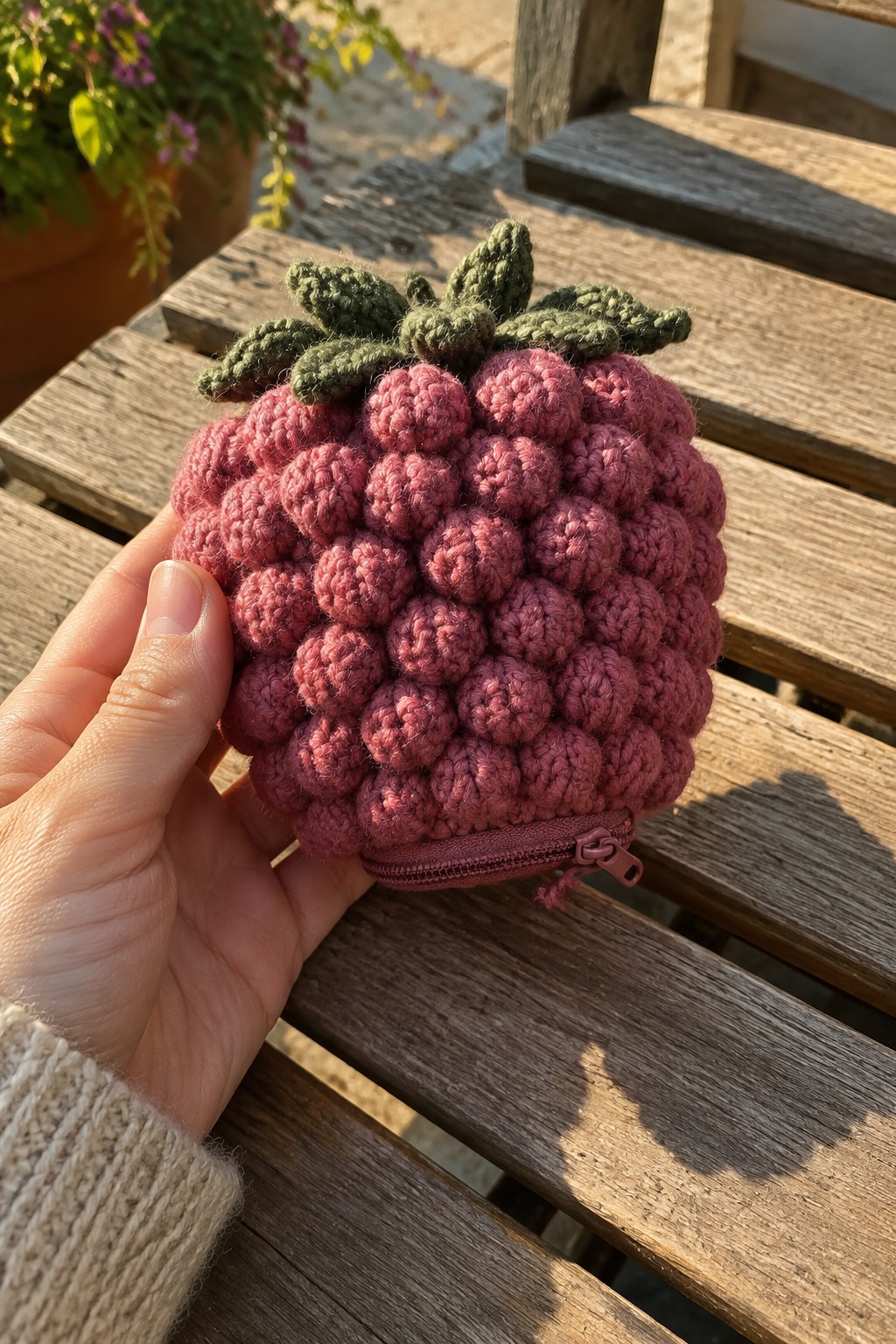 Hand in beige sweater holding crocheted pink raspberry pouch with green leaves on bench