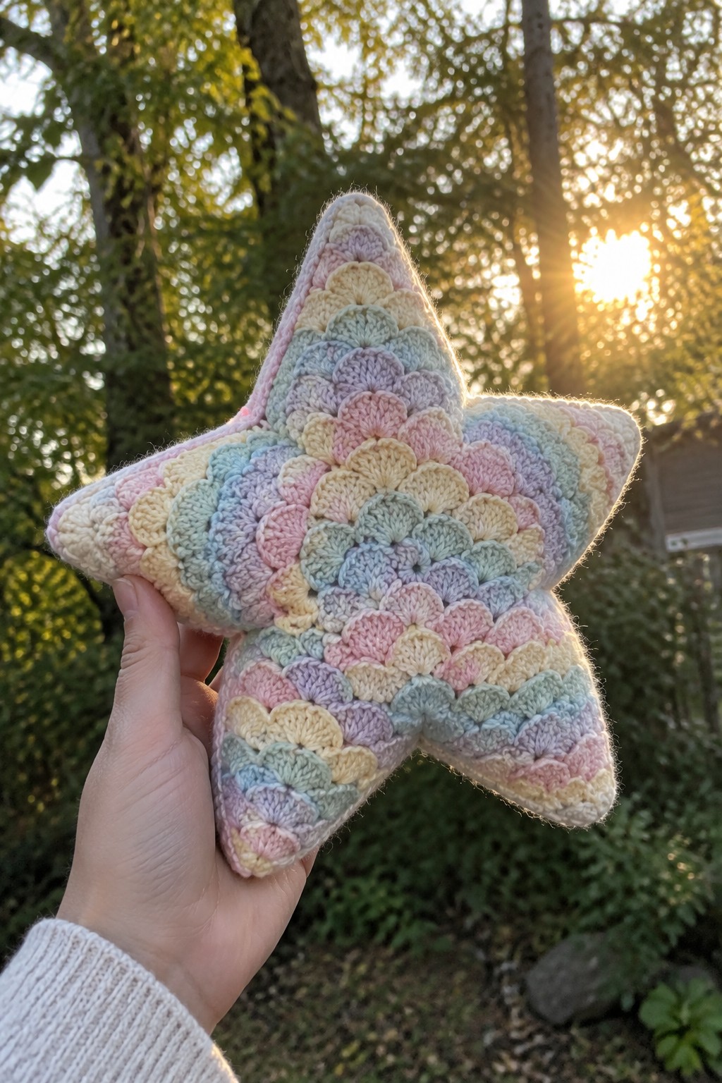 Hand holding pastel rainbow crocheted star pillow against sunny wooded backdrop