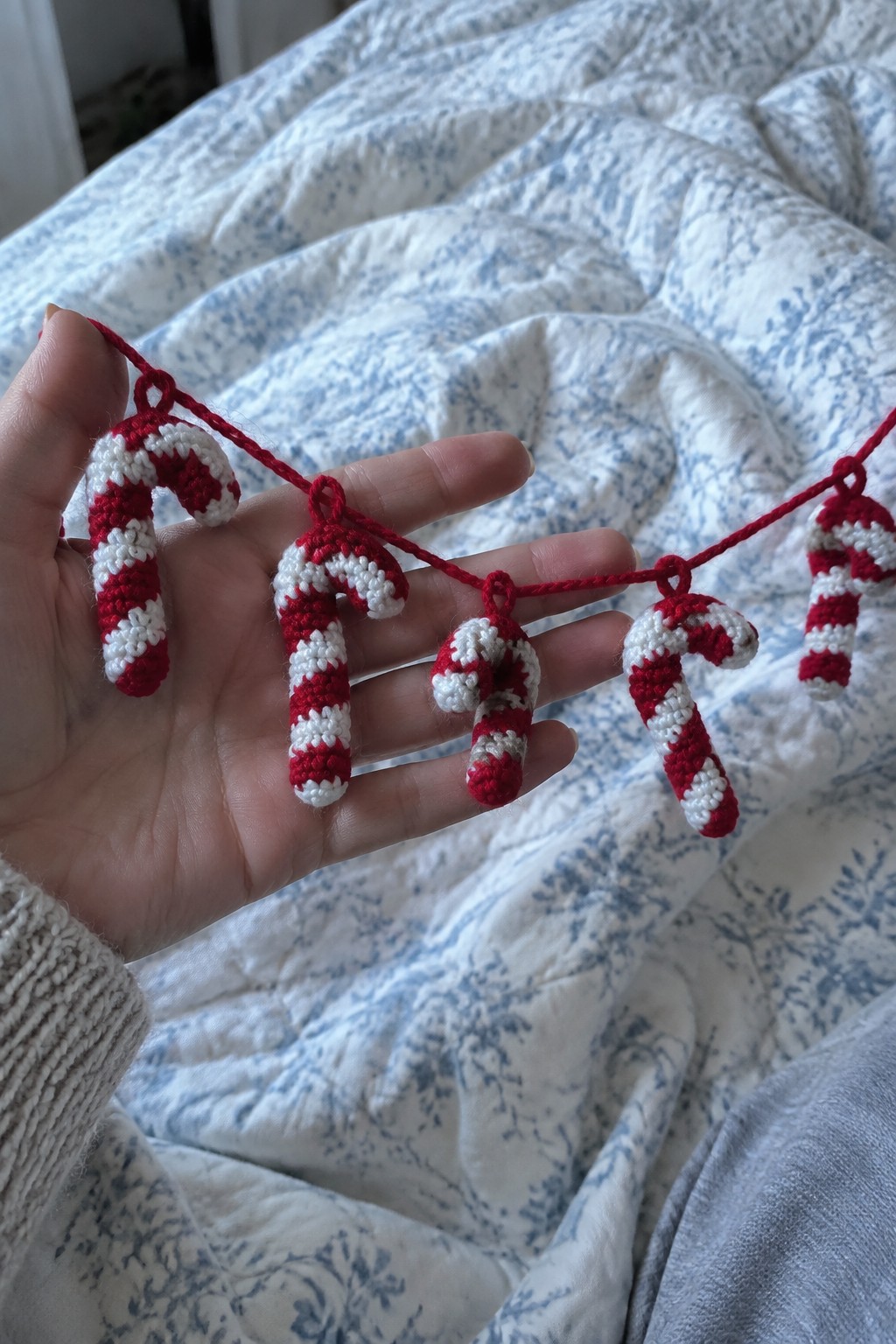 Hand holding five crocheted red-and-white candy cane ornaments on red string over quilt