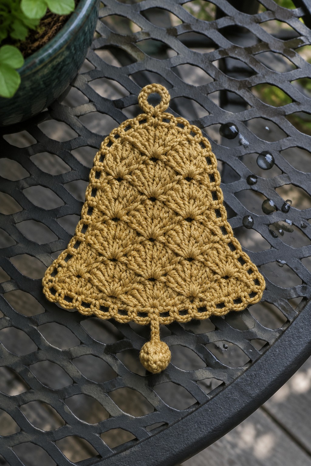 A mustard yellow crocheted bell with a top loop and bottom ball detail resting on a metal outdoor table.