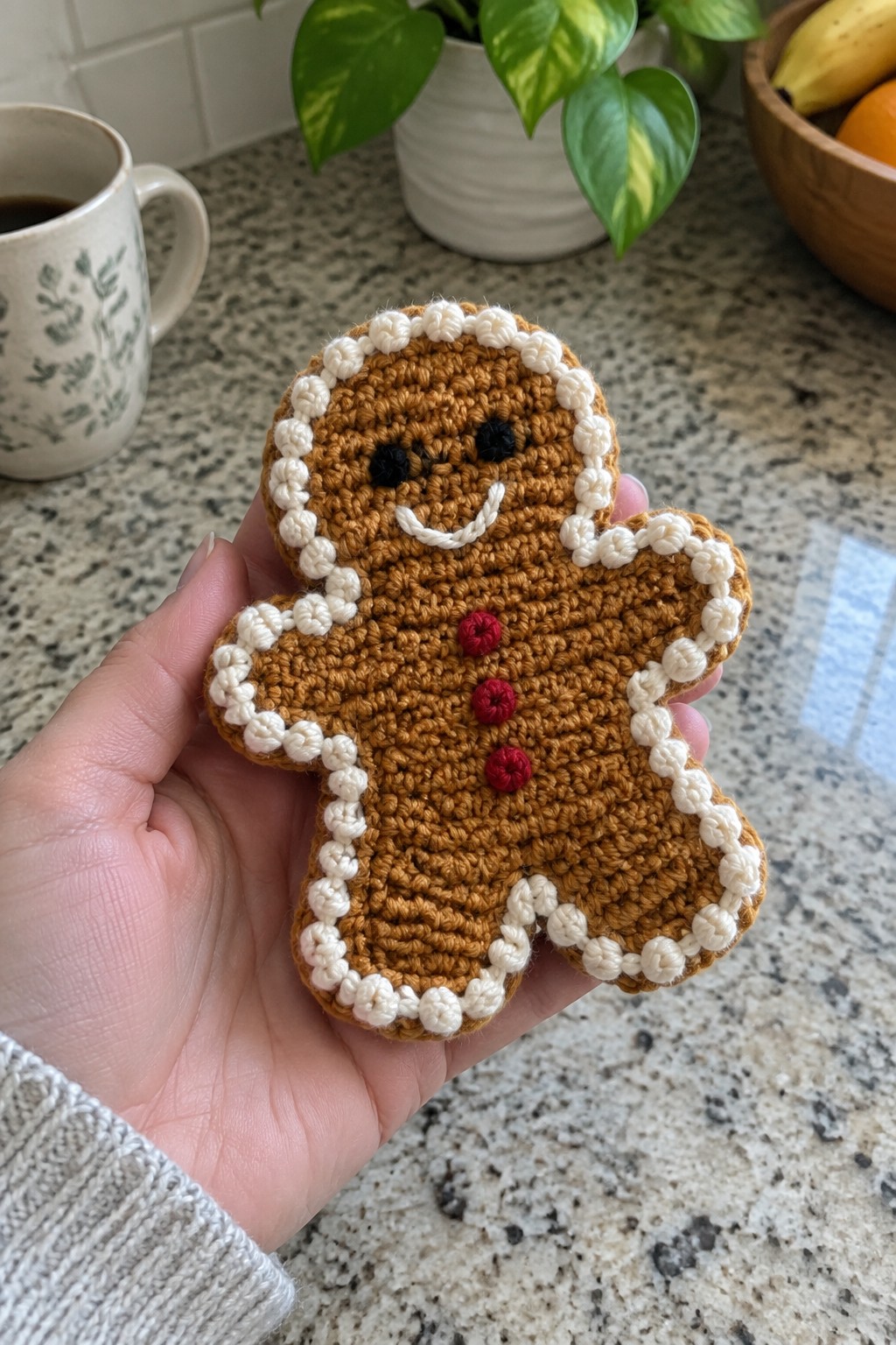 Hand holding crocheted gingerbread man with white trim and red buttons on counter
