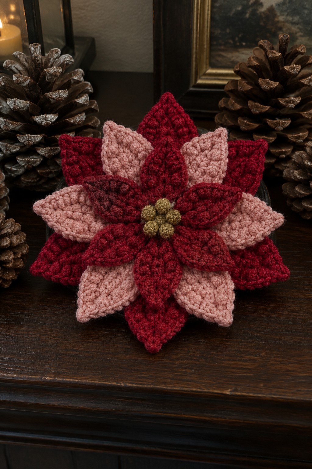 A crocheted poinsettia coaster in red and pink tones with a green center rests on a wooden surface next to pinecones.