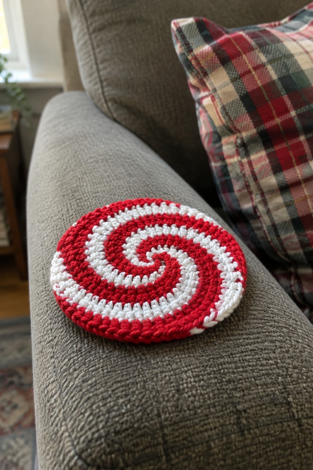Red and white crocheted spiral coaster on gray couch beside plaid pillow.