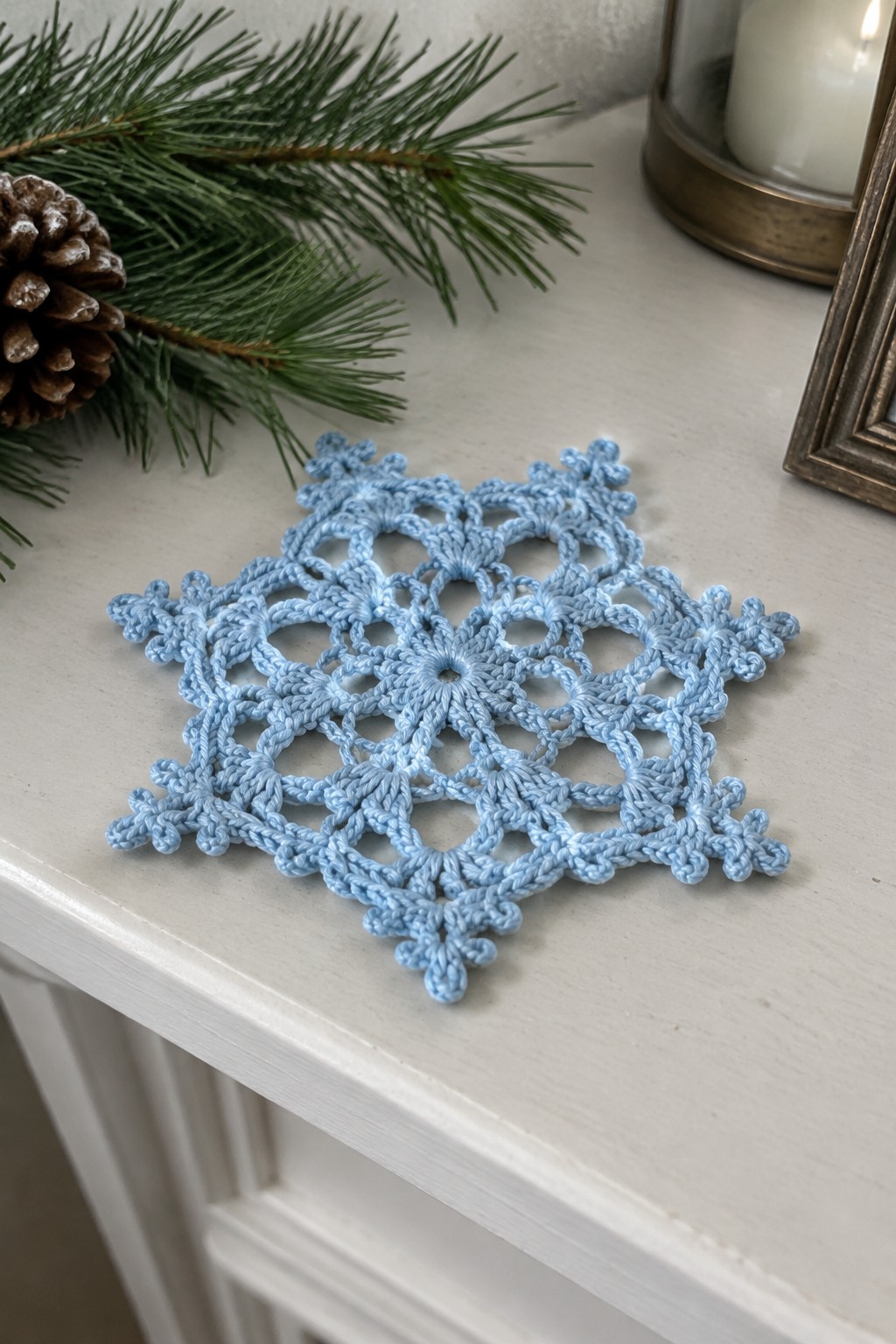 Light blue crocheted snowflake doily on white surface with pine branch and candle