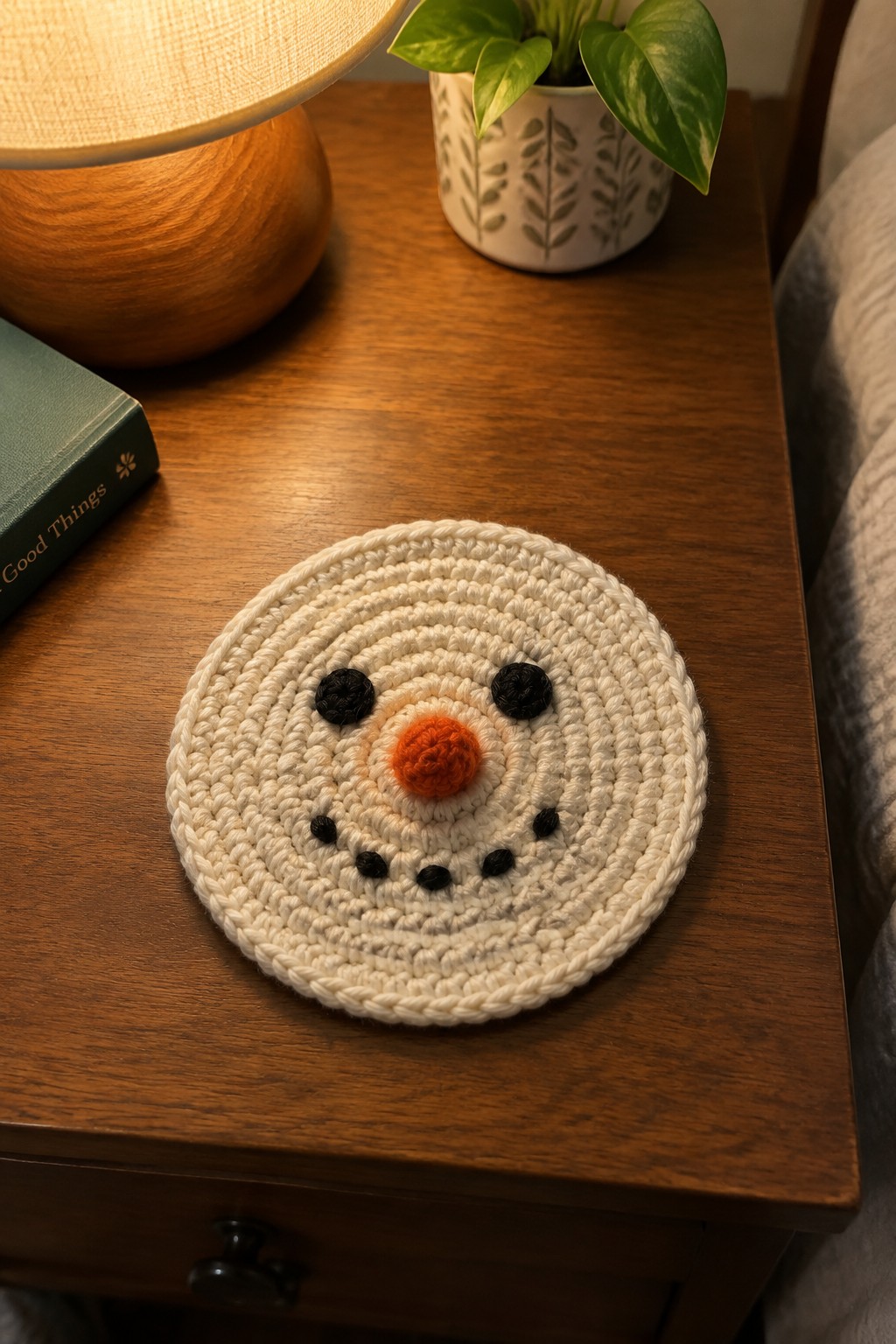 A round cream crochet coaster with a snowman face made from black and orange yarn rests on a wooden table.
