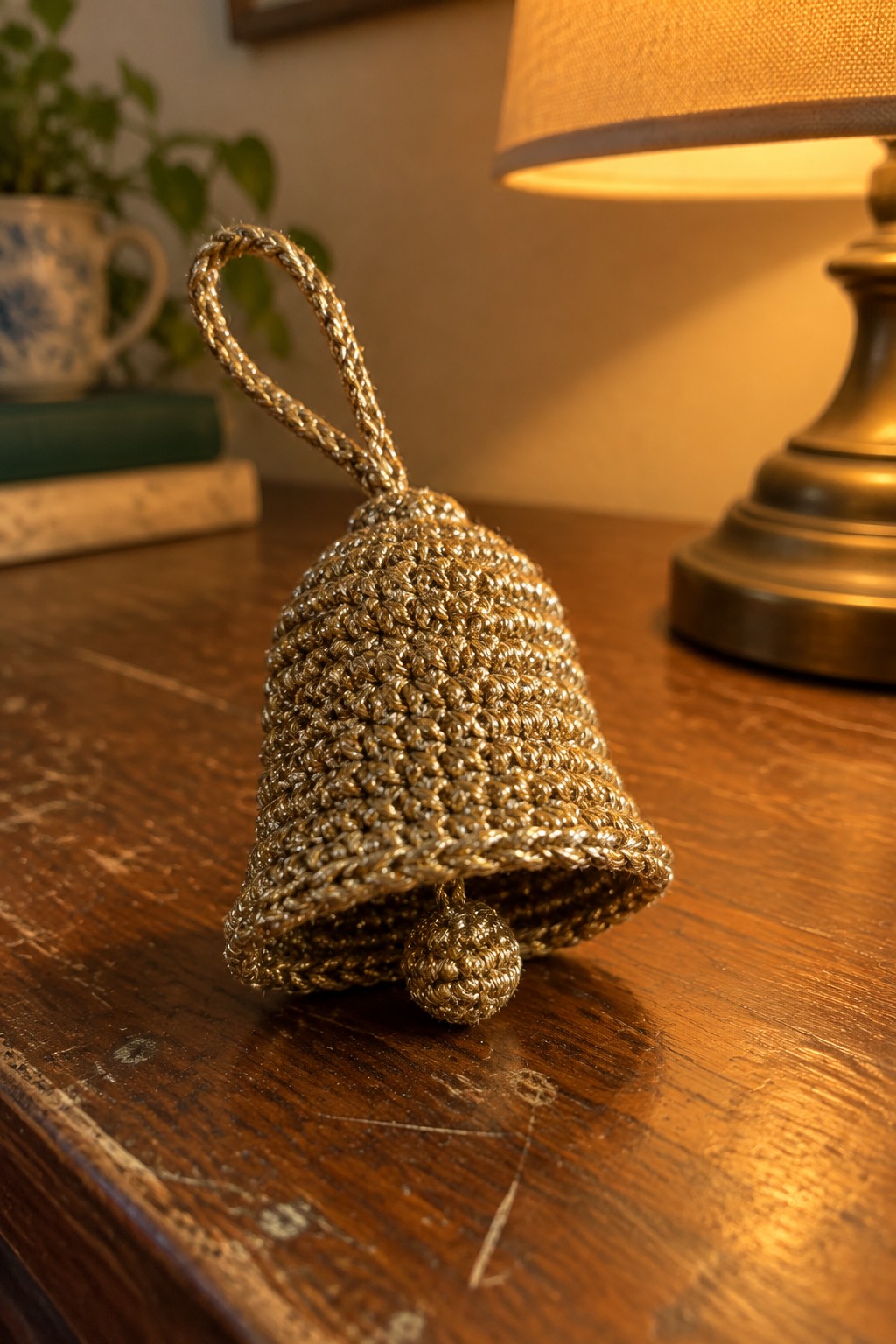 Crocheted golden bell ornament on wooden table with brass lamp nearby