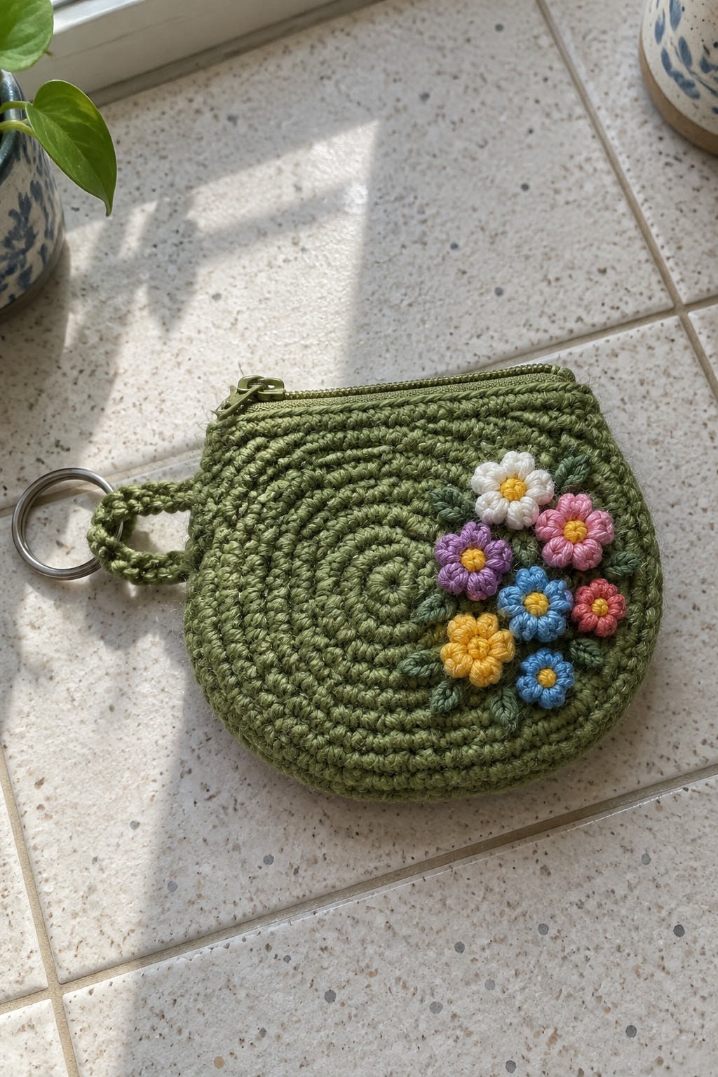 Green crocheted zippered pouch with colorful floral appliques and metal keyring on tiles