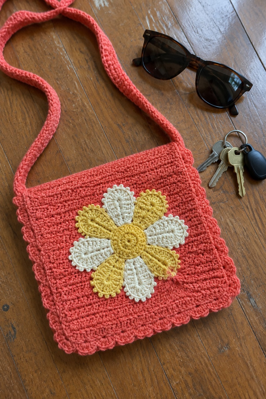 Crocheted pink bag with yellow-white daisy beside sunglasses and keys on wood.