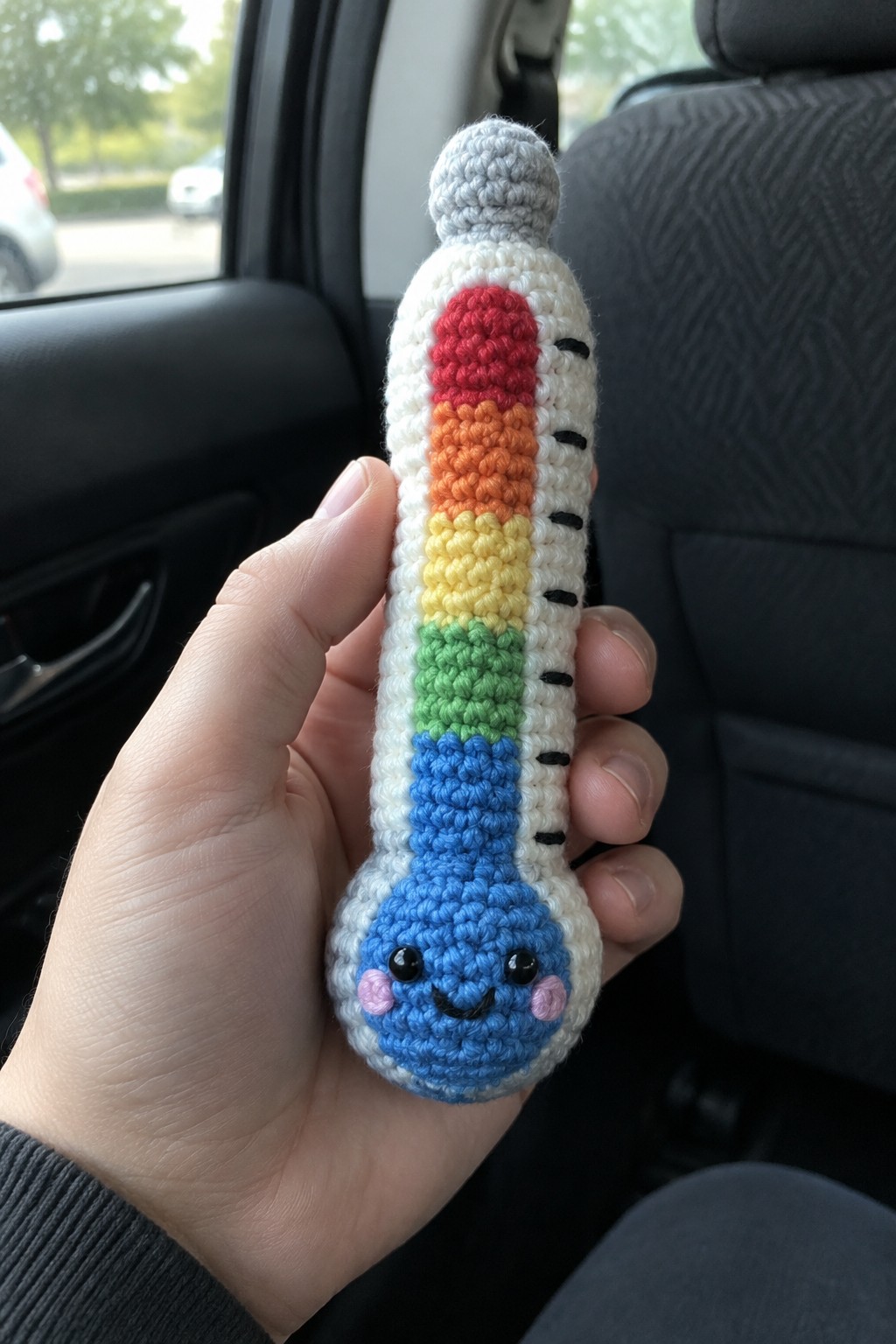 Hand holding crocheted rainbow thermometer plush toy with smiling face in car.