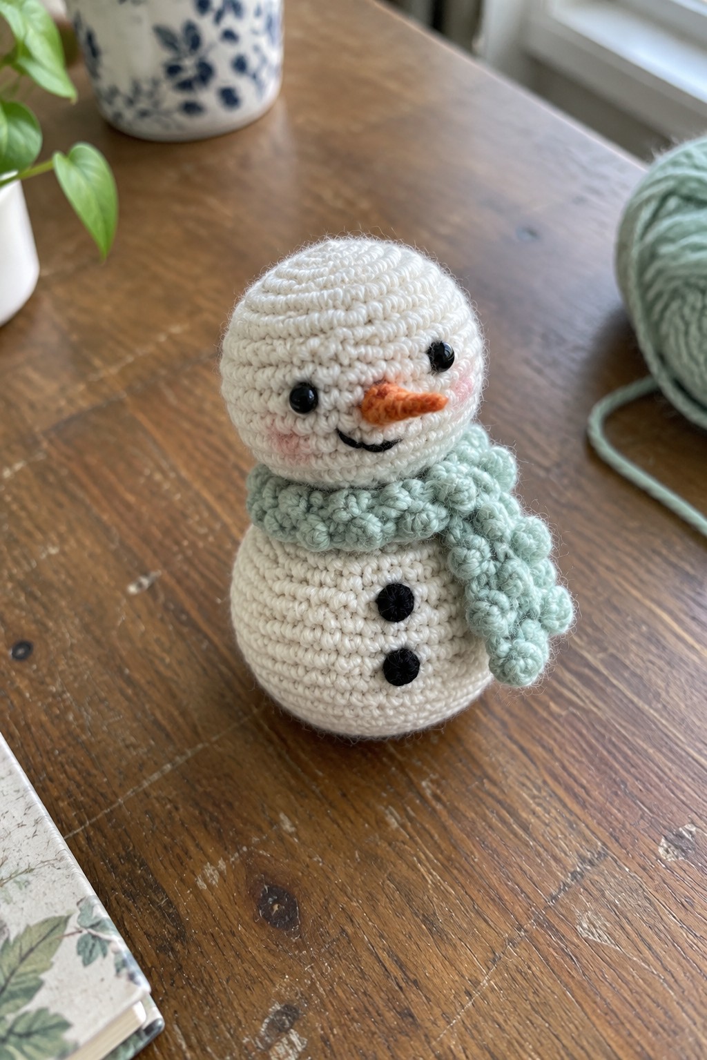 Crocheted white snowman with light green scarf, orange nose, and black buttons on wood.