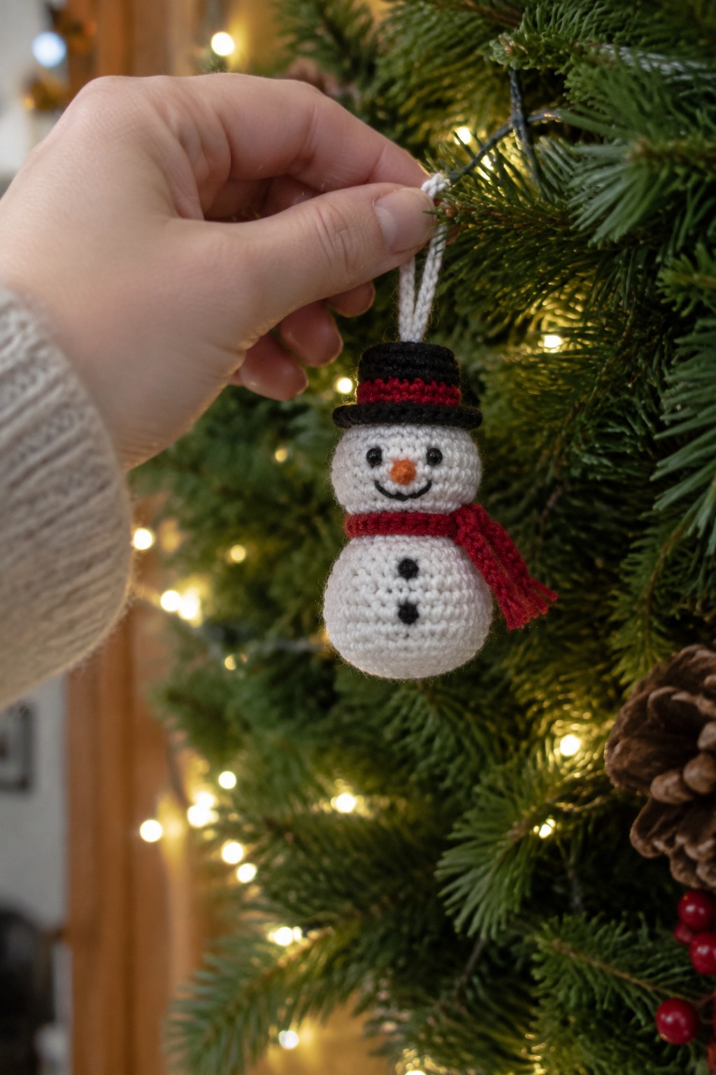 Hand holding crocheted snowman ornament with black hat and red scarf on lit tree.