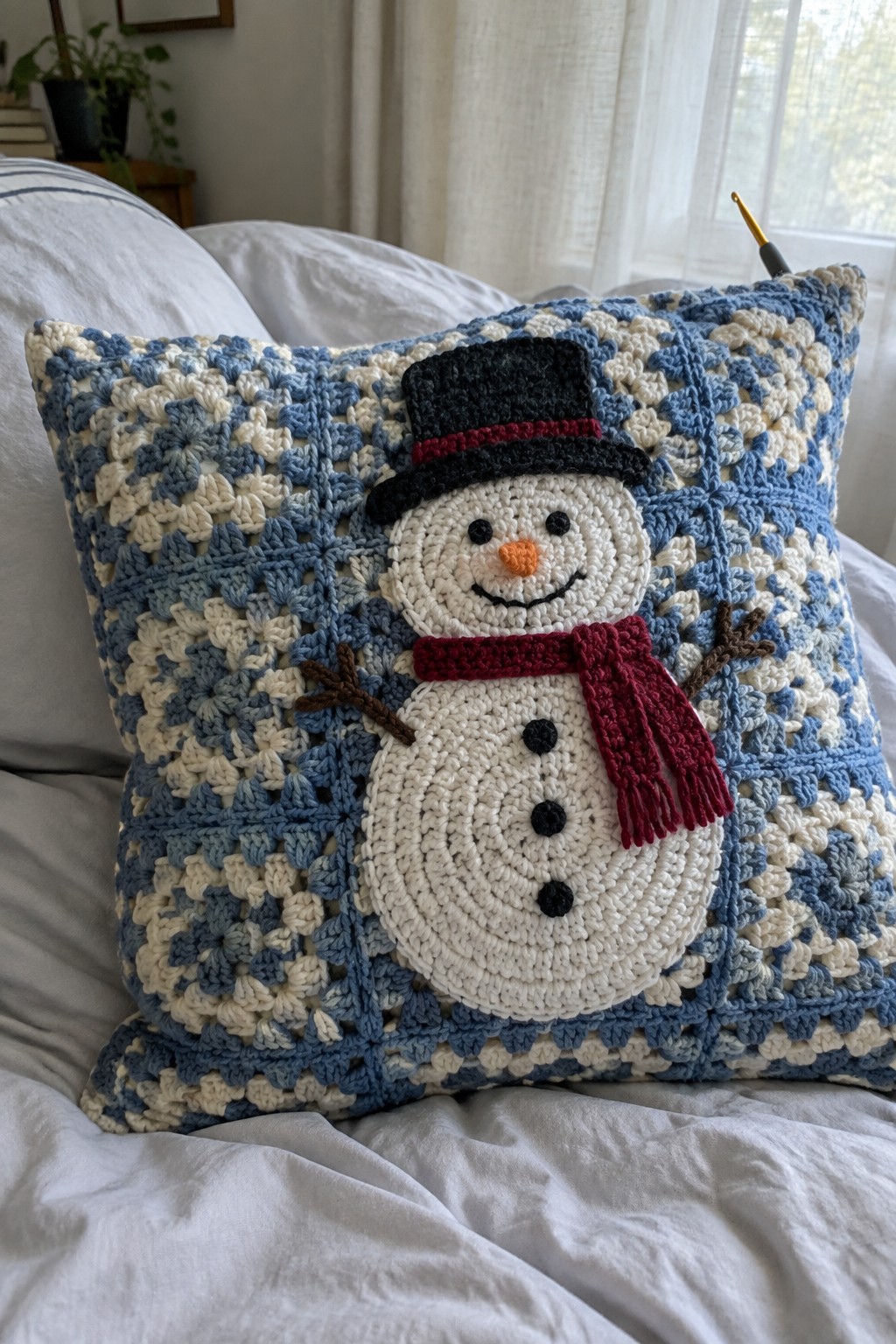 Blue and white crocheted pillow with snowman in black hat and red scarf on bed.