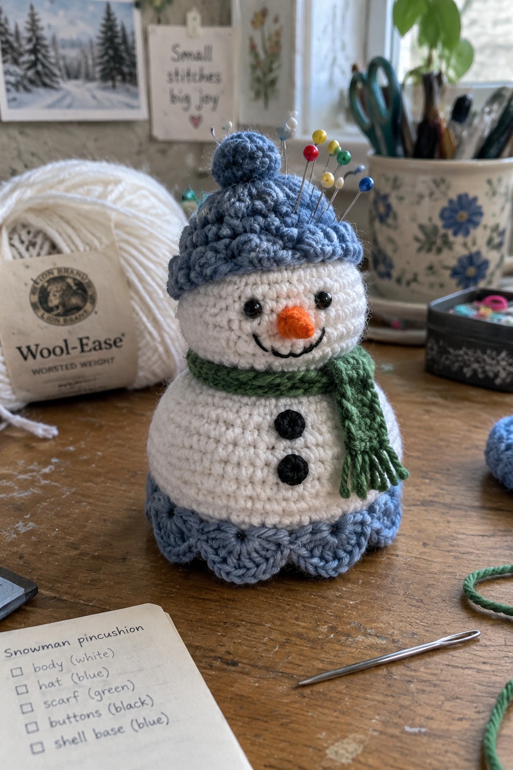 Crocheted snowman pincushion with blue hat, green scarf, and pins on wooden table