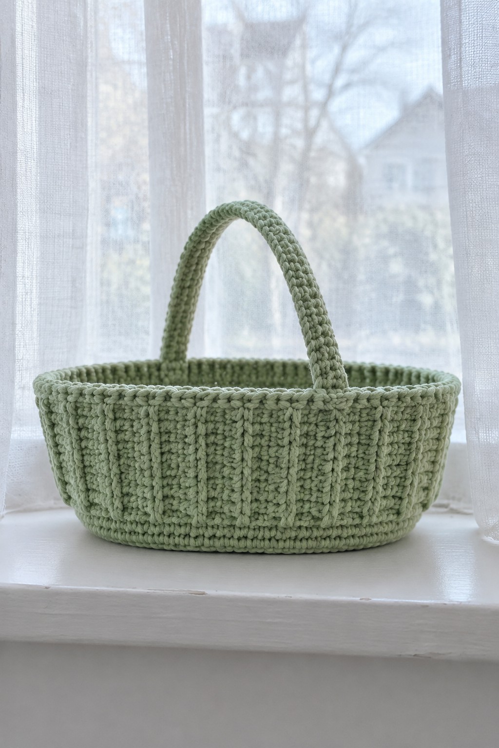 Light green crocheted basket with handle on white windowsill by sheer curtains
