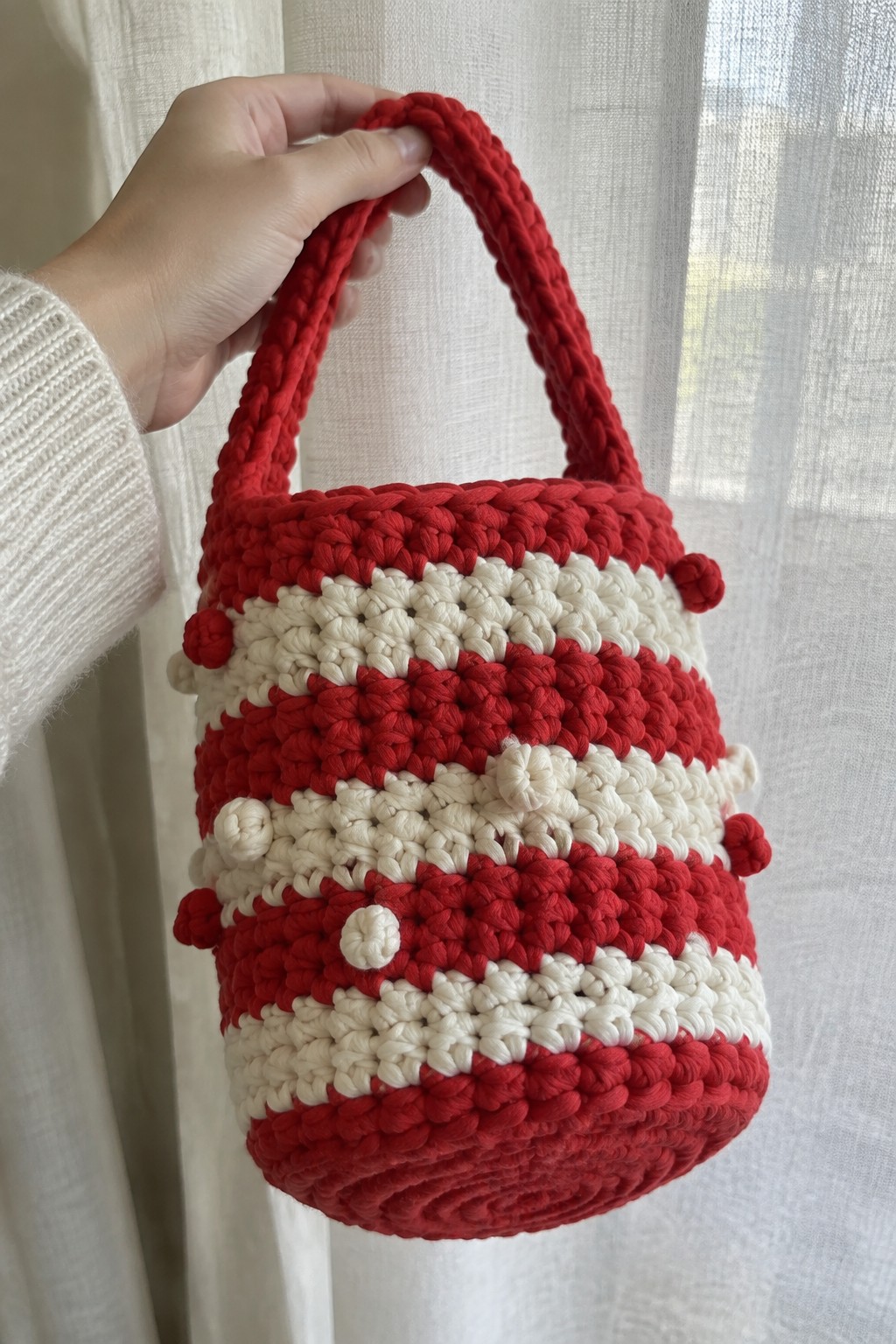 Hand in white sweater holding red and white striped crocheted bag with pom-poms