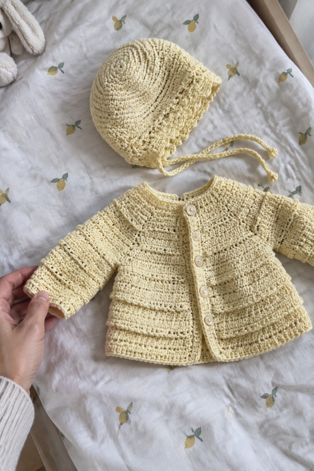 Yellow knitted baby cardigan and bonnet on lemon-patterned fabric, hand holding sleeve