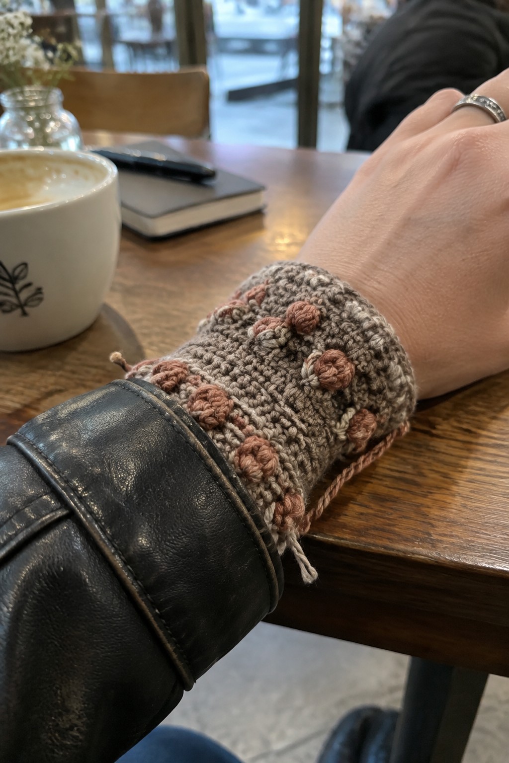 Crocheted wrist warmer with pink bobbles on arm in black leather jacket at wooden table