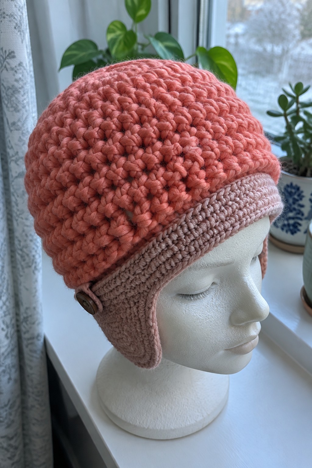 White mannequin wearing coral crocheted hat with light pink ear flaps and button