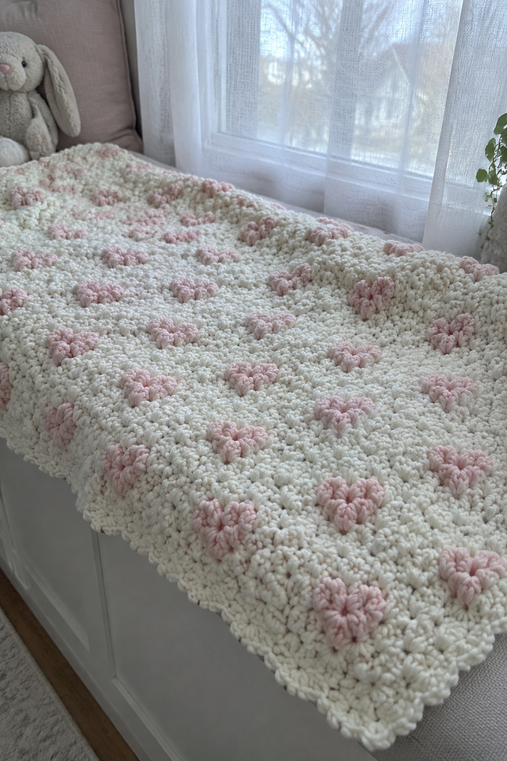 White crocheted blanket with pink heart patterns on a windowsill beside a stuffed bunny