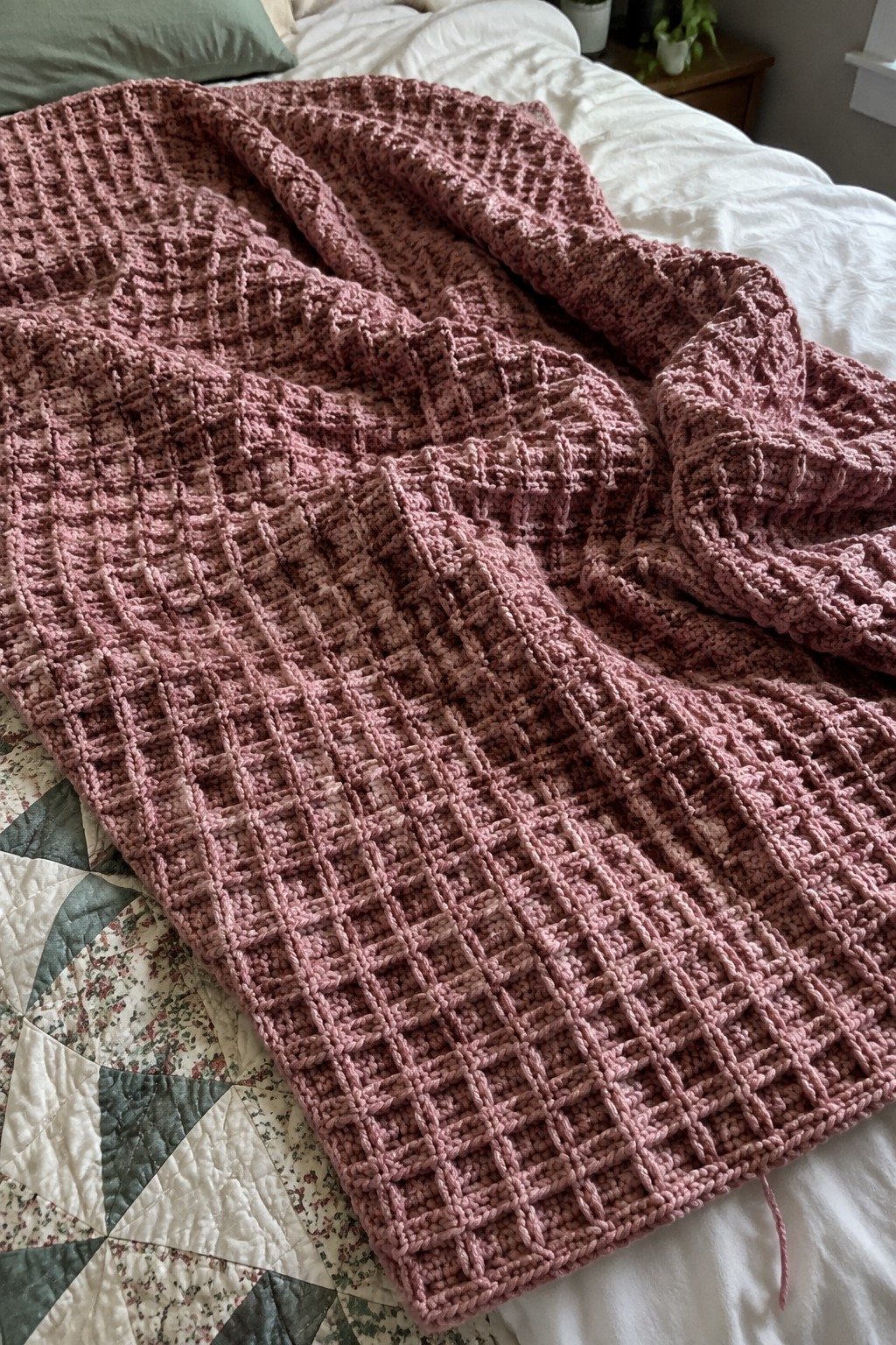 Pink crocheted waffle blanket draped over bed with patchwork quilt
