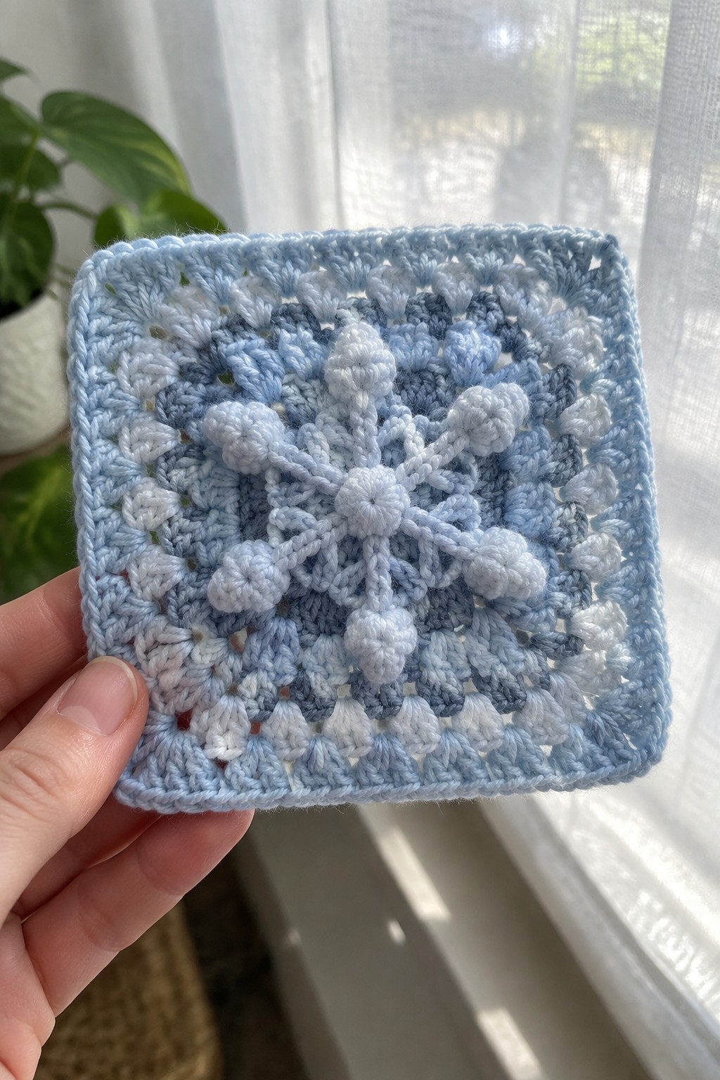 Hand holding light blue crocheted square with white snowflake pattern and bobbles