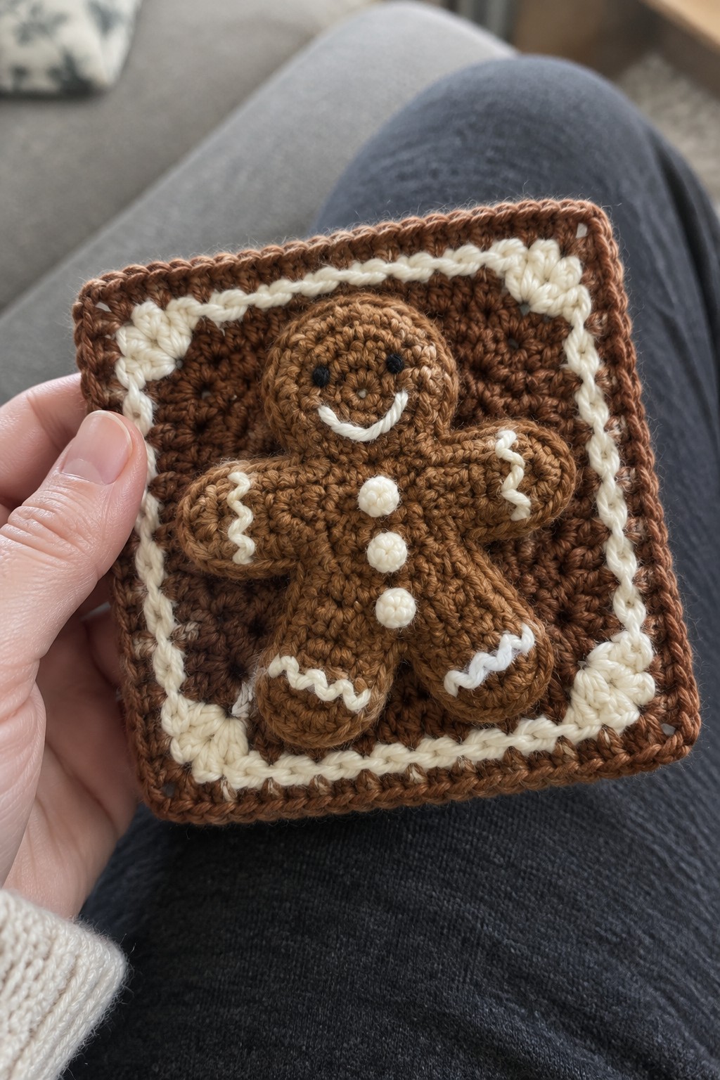 Hand holding brown crocheted granny square with white-iced gingerbread man on blue jeans.